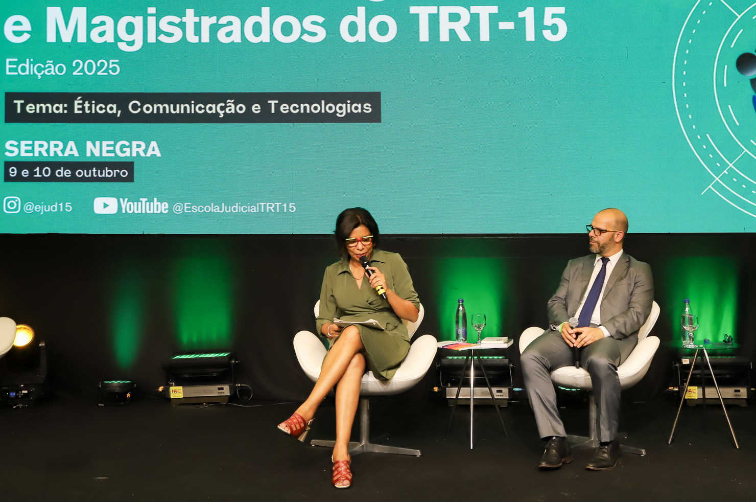 "Dra. Rosilene está sentada numa cadeira, no palco, enquanto segura um papel e fala ao microfone, sendo observada pelo escritor Oscar Krost"