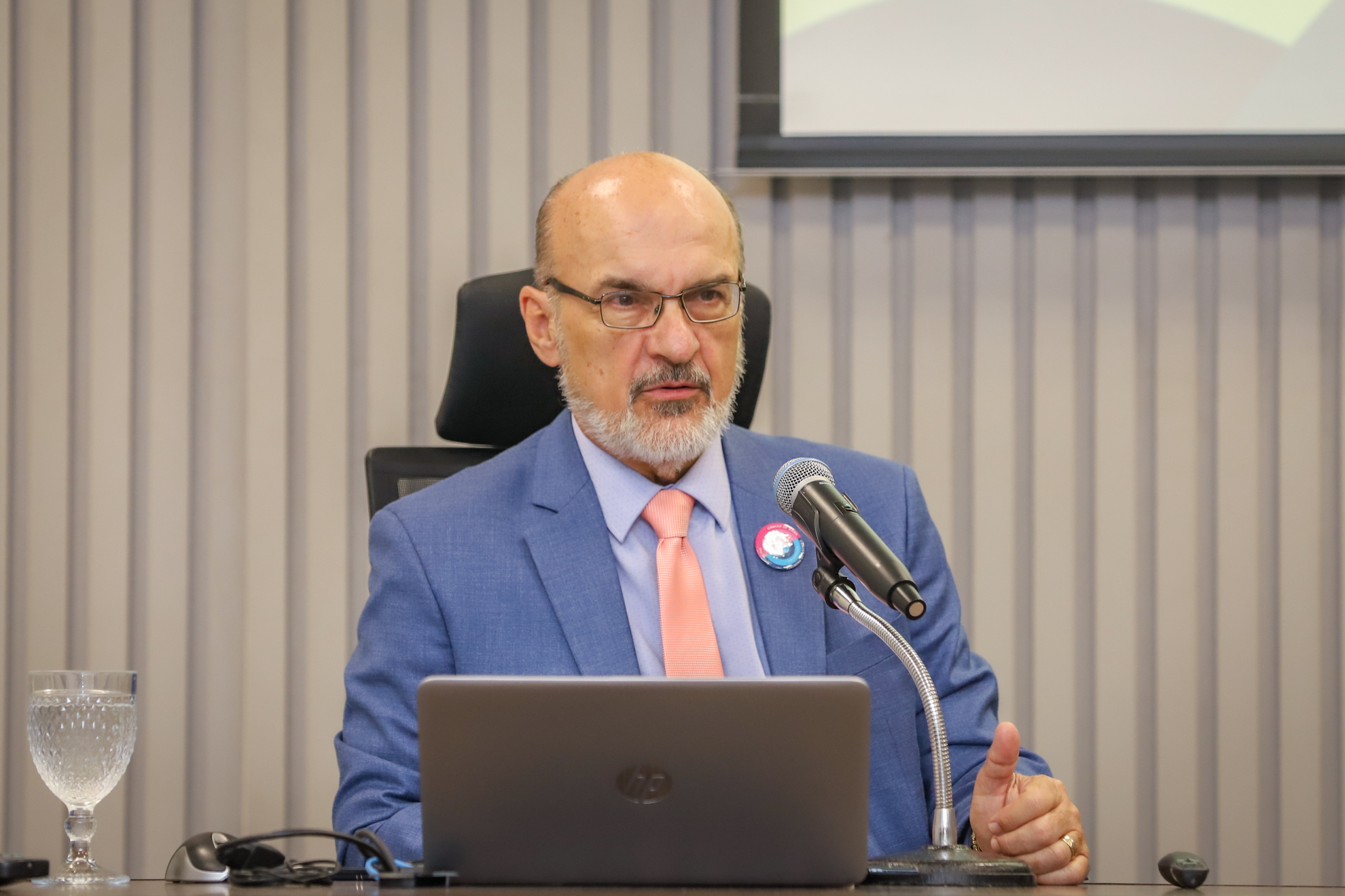 "Dr. Edmundo fala ao microfone, sentado à mesa de trabalhos"