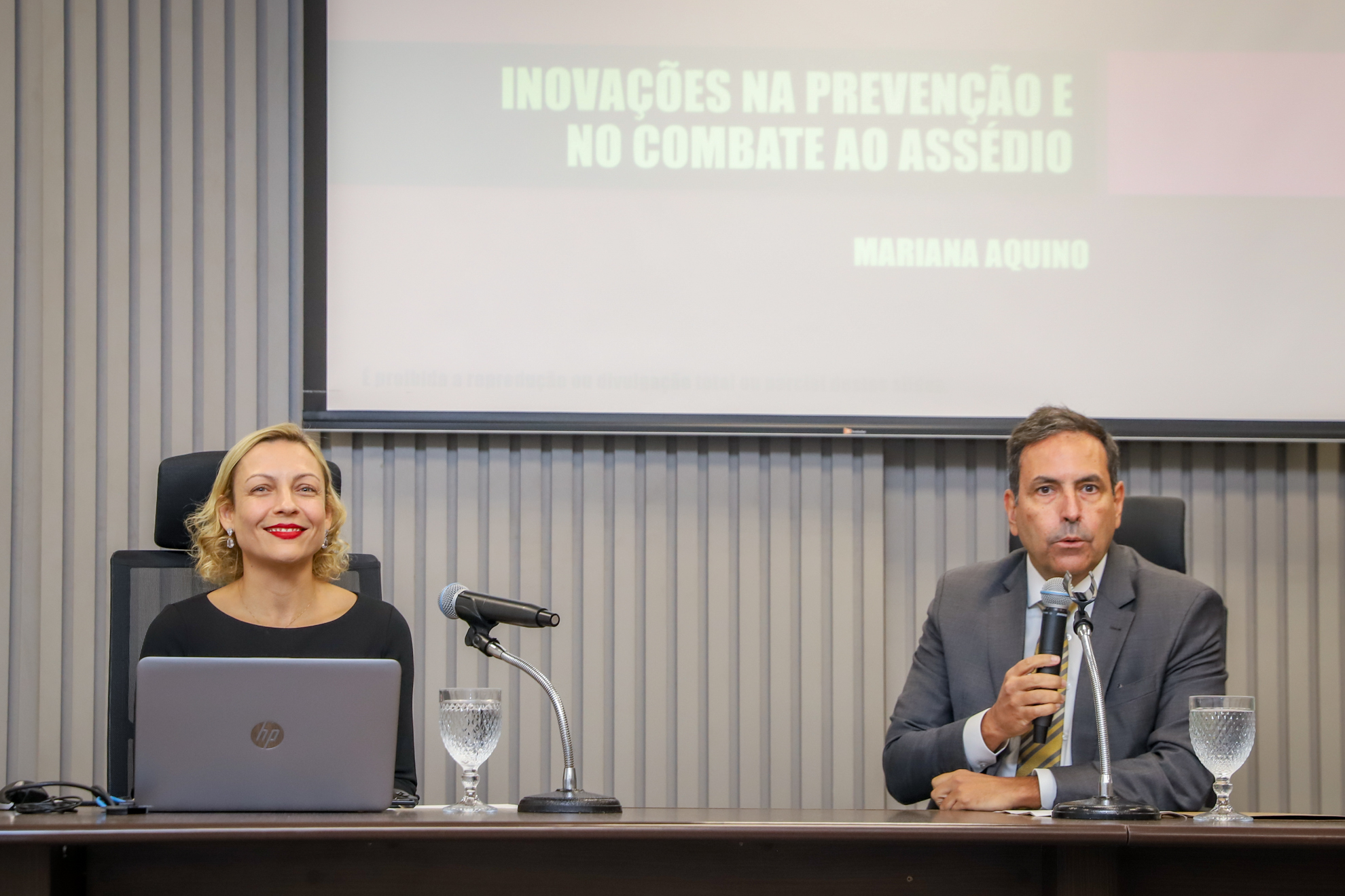 "Dr. Marcos Porto fala ao microfone ao lado da palestrante, dra. Marina"
