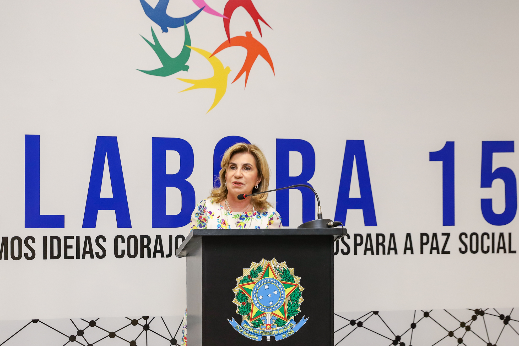 "Dra. Ana Paula fala ao microfone, no púlpito, em frente a um painel com o logotipo do Co.Labora 15"