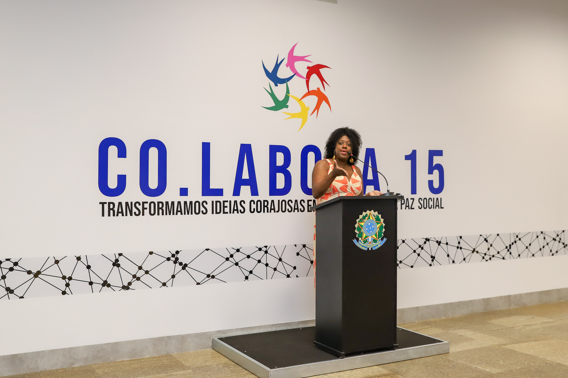 "Eliane fala ao microfone, no púlpito, em frente a um painel com o logotipo do Co.Labora 15"