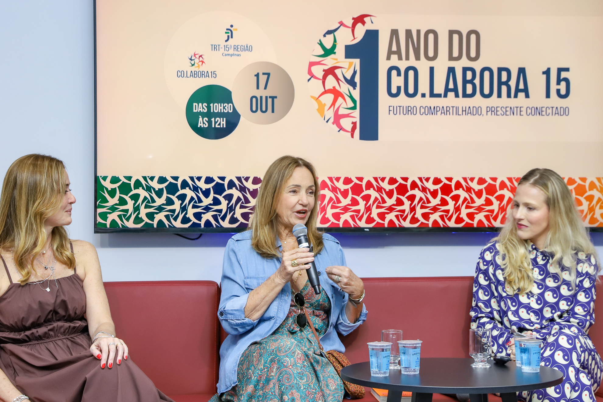 "Dra. Gisela segura o microfone ao lado de Dra. Daniela e Michelle. Elas estão sentadas em um sofá vermelho, no palco do auditório em frente a um telão"