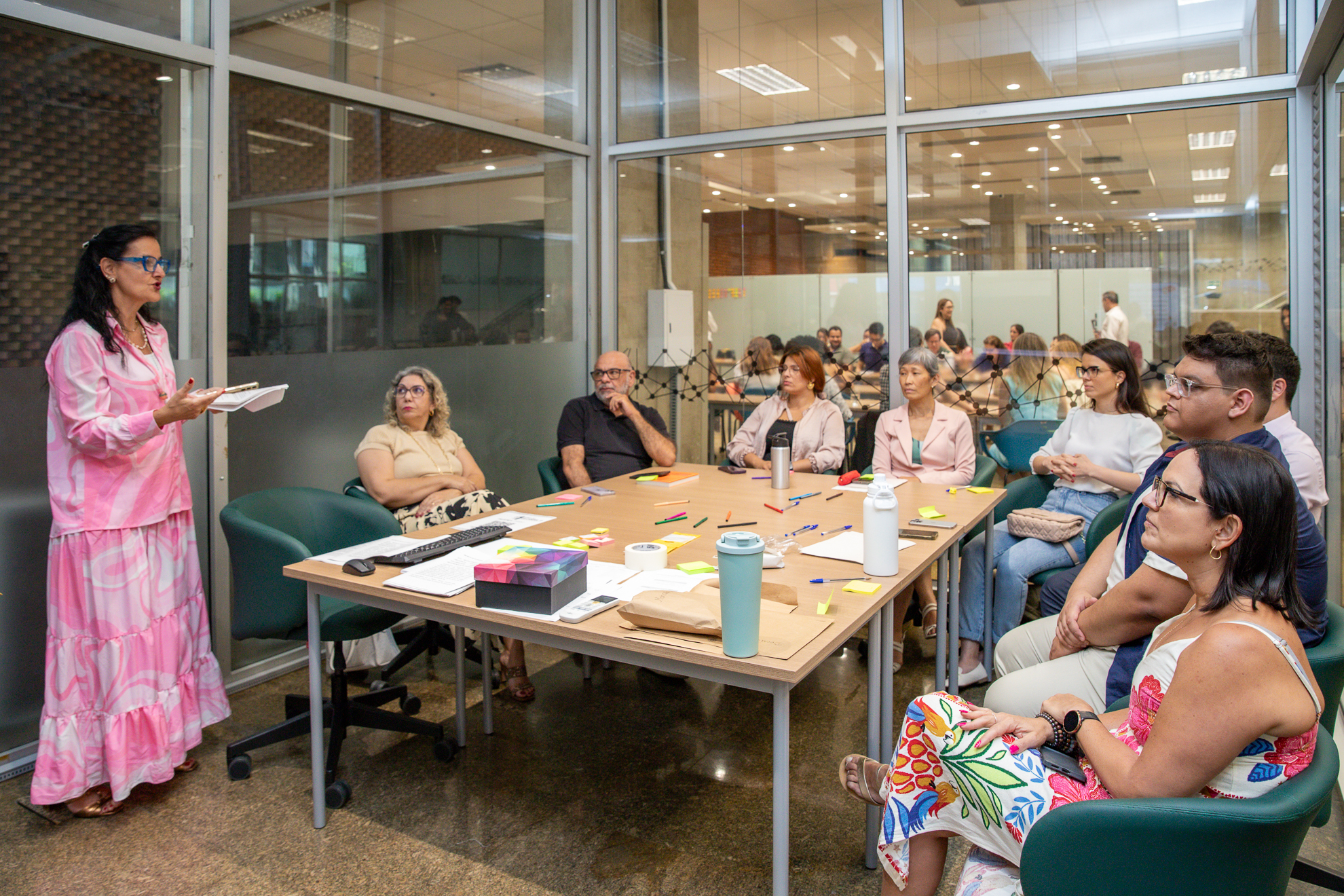 "Juíza Juliana comanda oficina sobre linguagem simples, sendo observada pelos participantes que estão sentados à mesa"