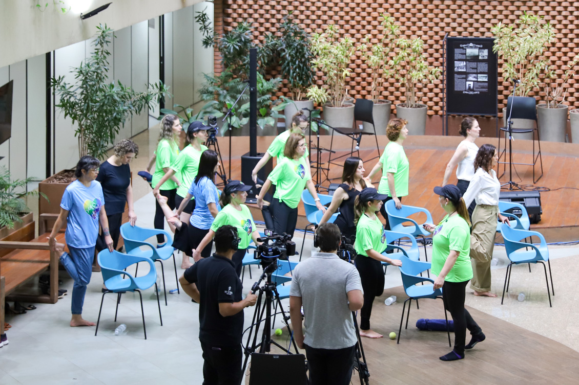"Servidores fazem exercício de pilates utilizando cadeiras azuis. Em primeiro plano, uma equipe faz a filmagem para transmissão"