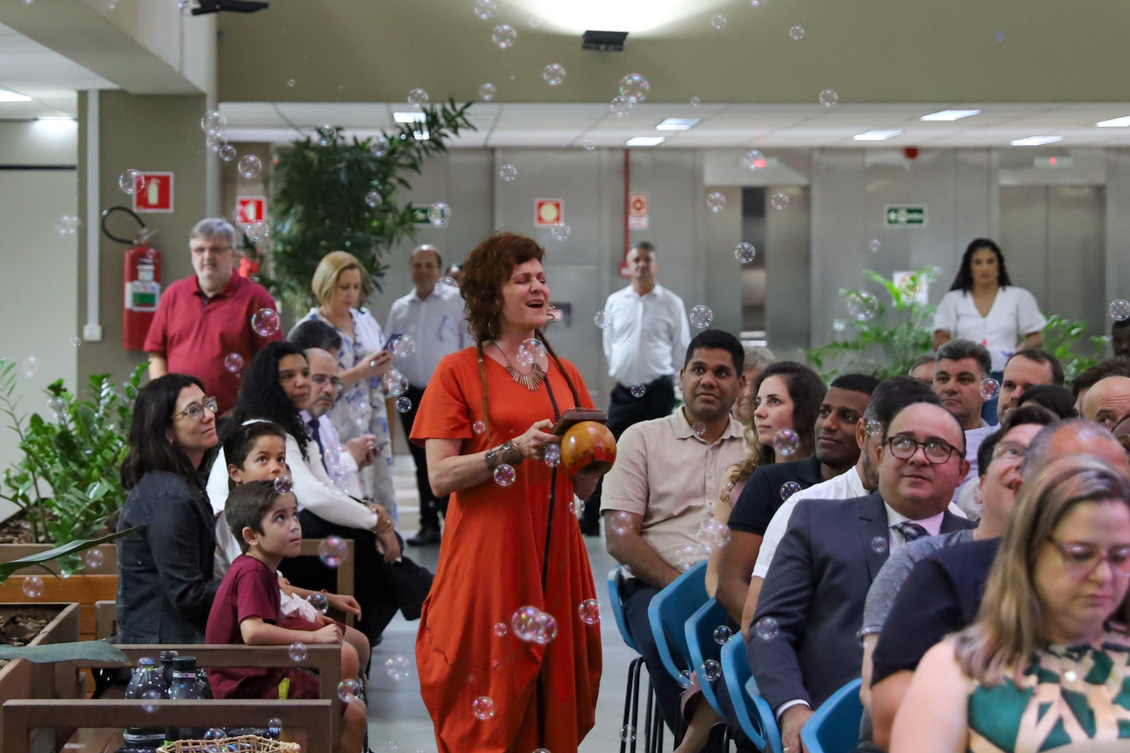 "Ana Person canta e toca um instrumento, sendo observada por servidores que estão sentados em cadeiras. Há bolas de sabão caindo sobre eles"