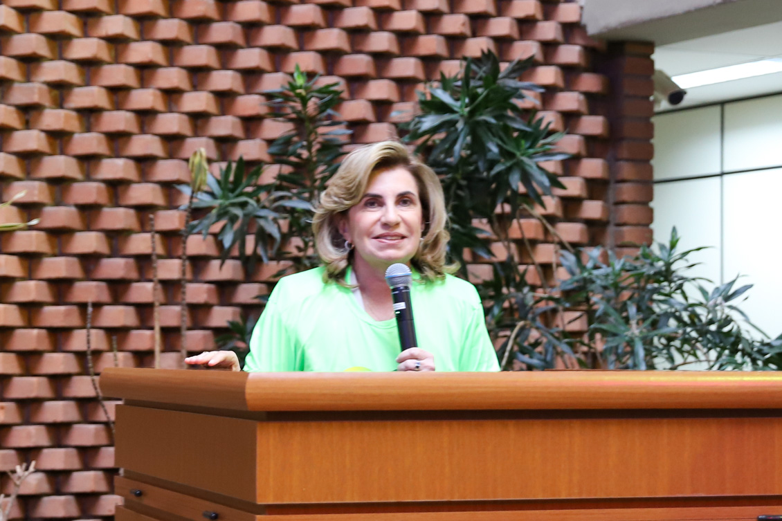 "Vestida com a camiseta da Semana do Servidor, Dra. Ana Paula fala ao microfone no púlpito de madeira"