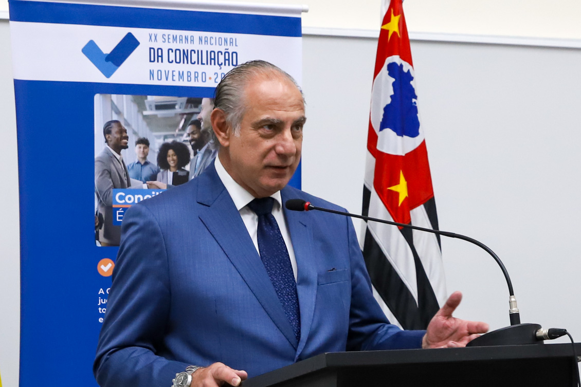 "Desembargador Fábio Aguiar fala ao microfone no púlpito de madeira. Ao fundo banner alusivo à campanha e a bandeira do Estado de São Paulo"