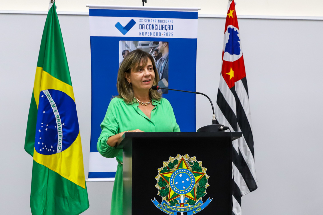 "Procuradora Alvamari fala ao microfone no púlpito de madeira. Ao fundo, banner alusivo à campanha, entre bandeiras do Brasil e do Estado de São Paulo"