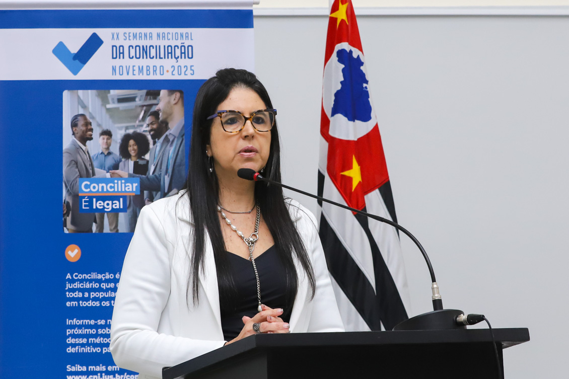 "Desembargadora Ana Cláudia fala ao microfone no púlpito de madeira. Ao fundo banner alusivo à campanha e a bandeira do Estado de São Paulo"