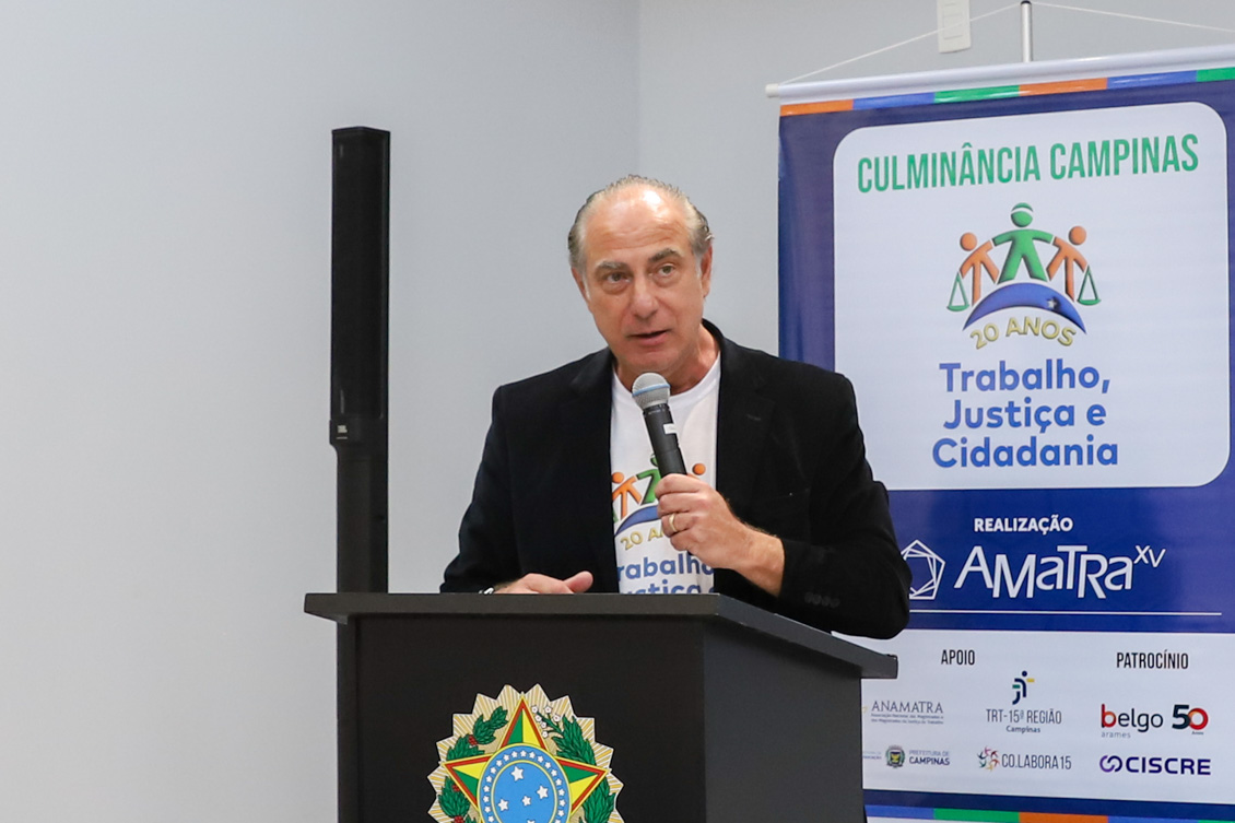 "Dr. Fábio fala ao microfone, no púlpito, em frente ao banner azul alusivo ao projeto"