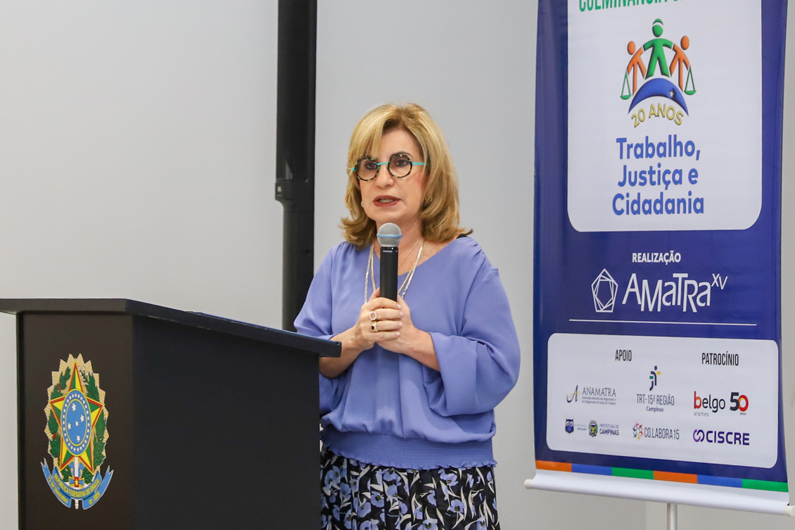 "Dra. Ana Paula está de pé, atrás do púlpito, falando ao microfone. Ao fundo, banner do projeto em azul"