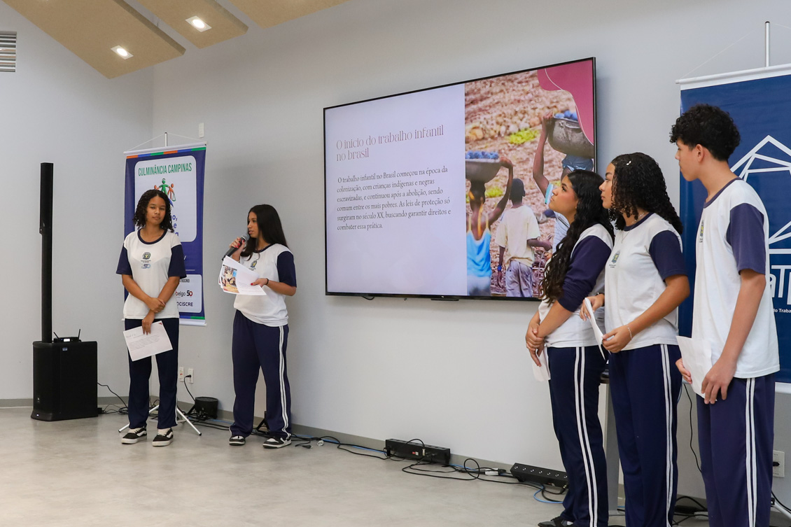 "Aluna está de pé, falando ao microfone, sendo observada por três outros alunos. No telão, informações sobre trabalho infantil"