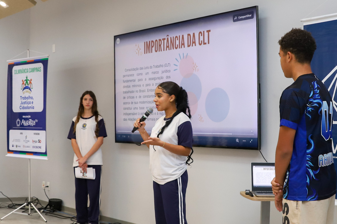 "Uma aluna está de pé falando ao microfone, com dois outros alunos observando. No telão, o tema: a importância da CLT"