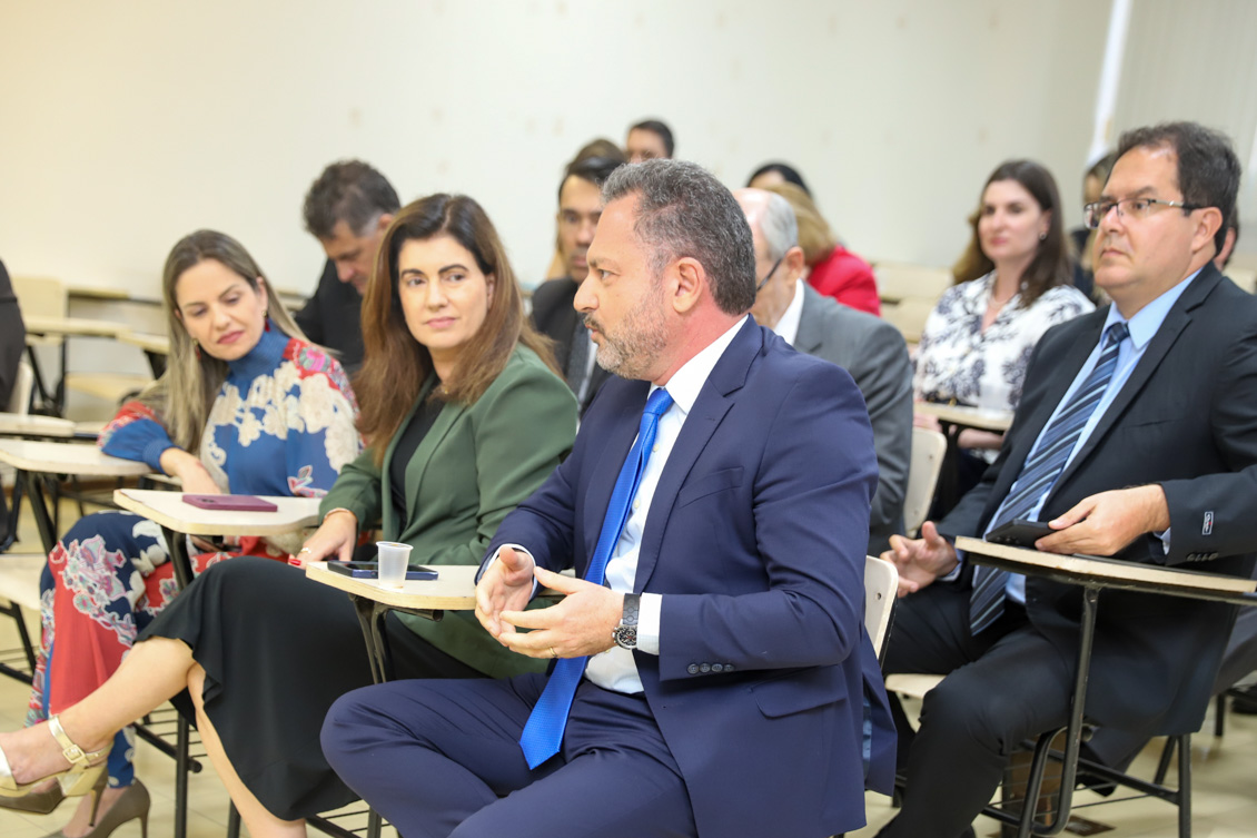 "Dr. Marcelo gesticula, observado pelos colegas. Todos sentados"