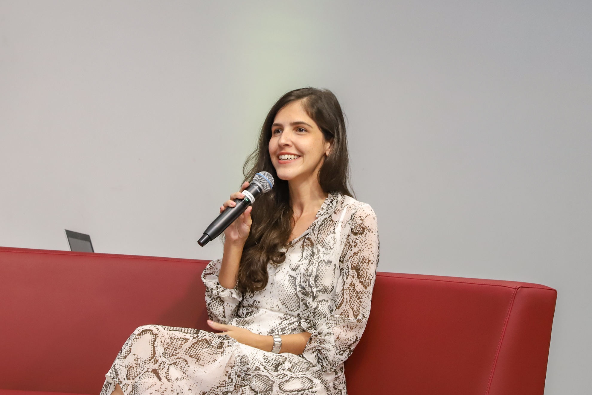 "Dra. Helena está sentada em um sofá vermelho, falando ao microfone"