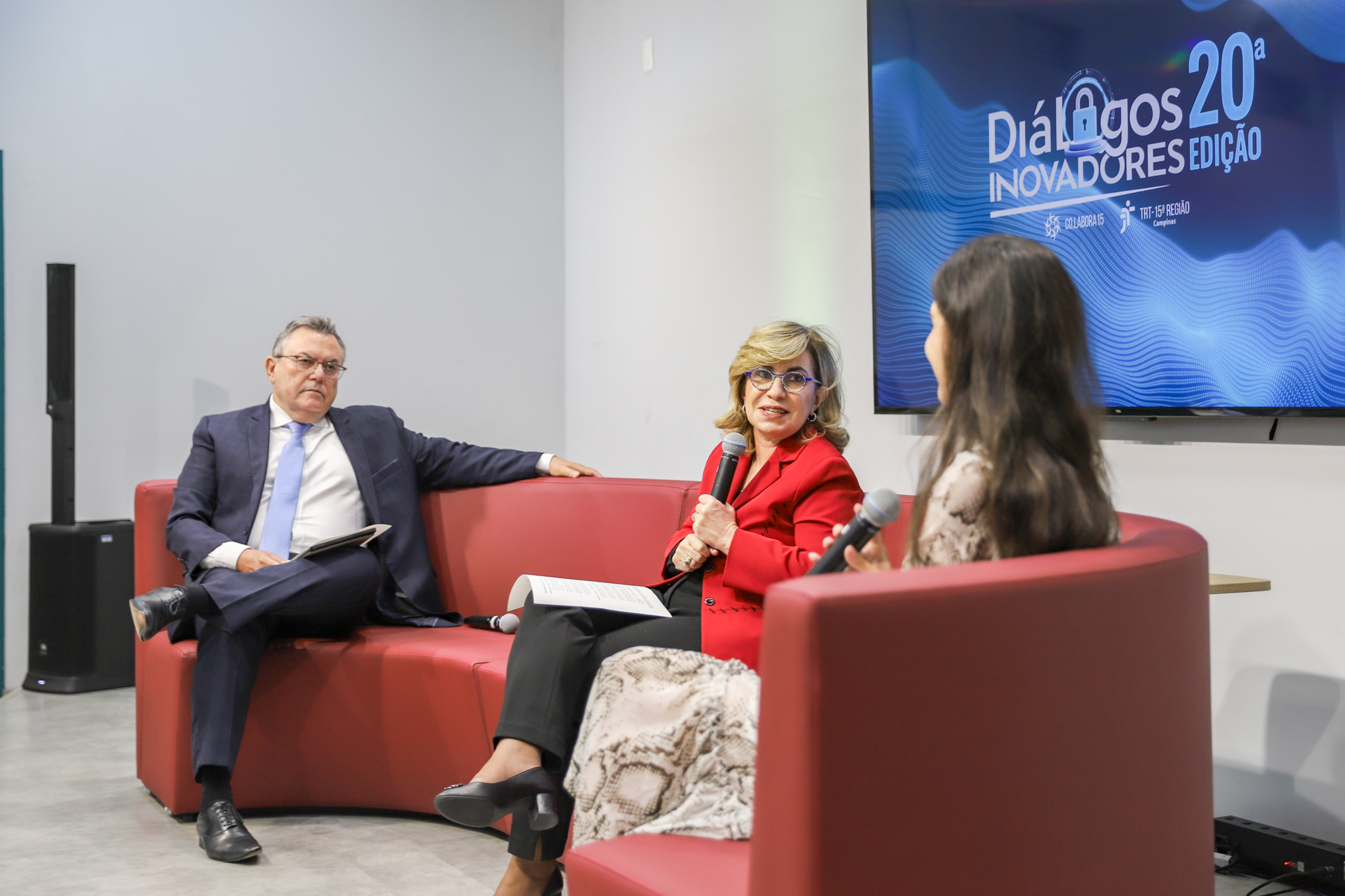 "Dr. Levi, Dra. Ana Paula com microfone na mão, e Dra. Helena estão sentados no sofá vermelho"