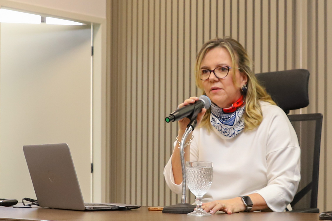 "Dra. Eleonora está na mesa alta, falando ao microfone, com um notebook do seu lado direito"