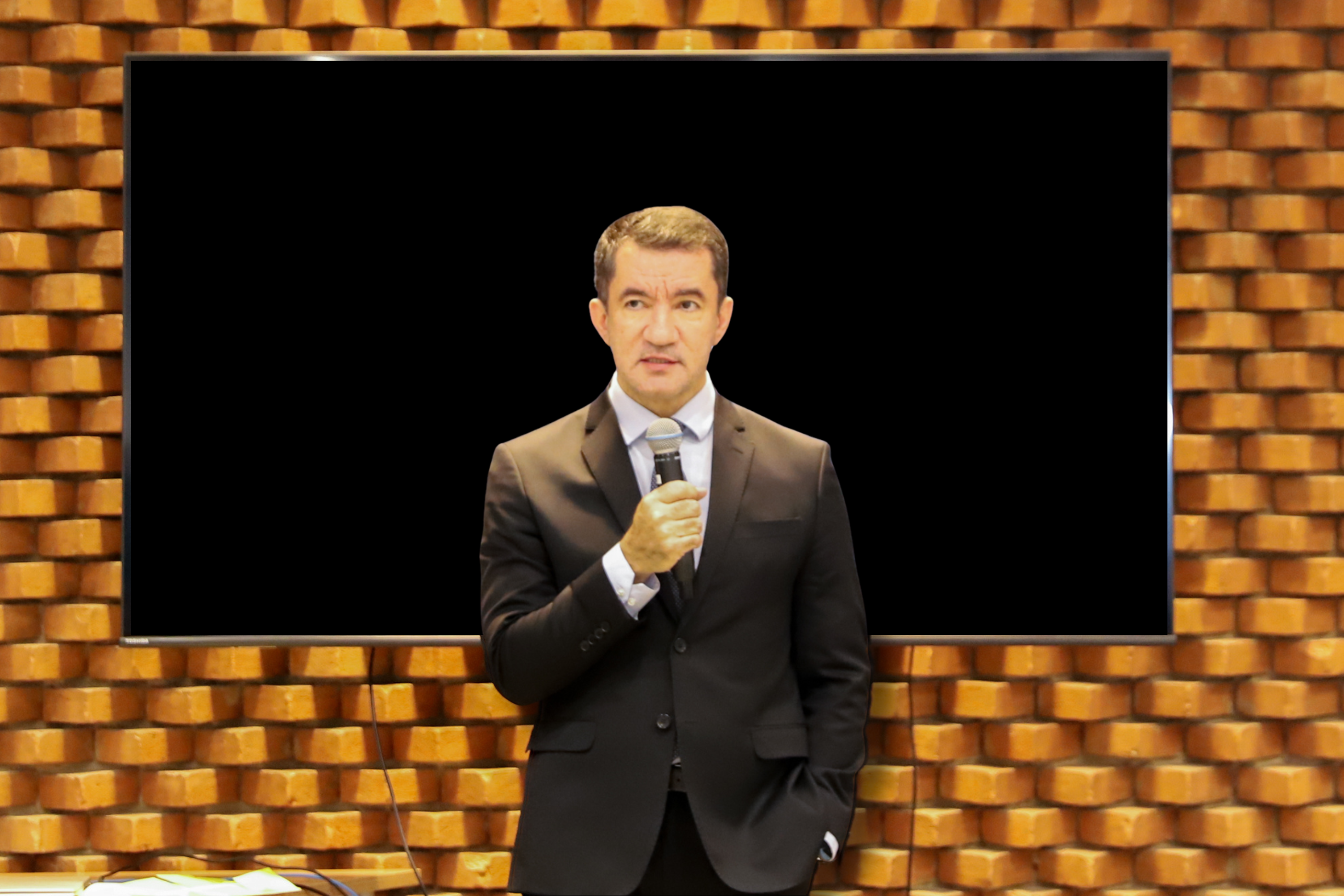 "Desembargador Renan fala ao microfone em frente a um telão de fundo preto"