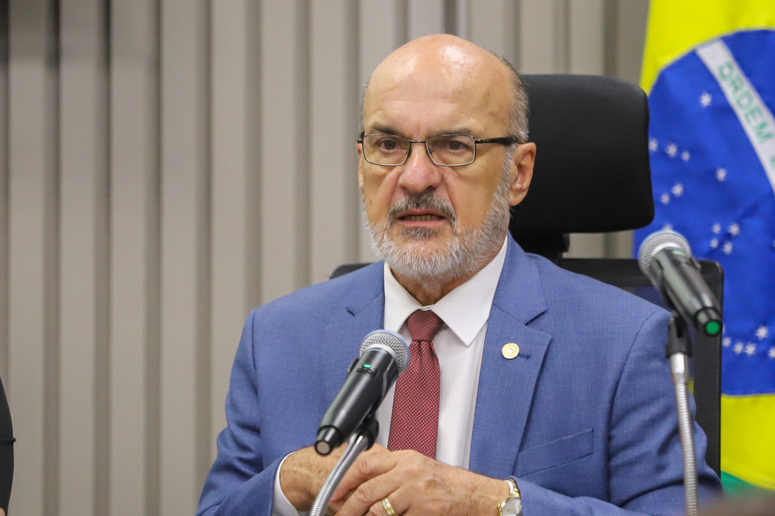 "Dr. Edmundo está sentado em mesa alta, falando ao microfone com a bandeira do Brasil ao fundo"