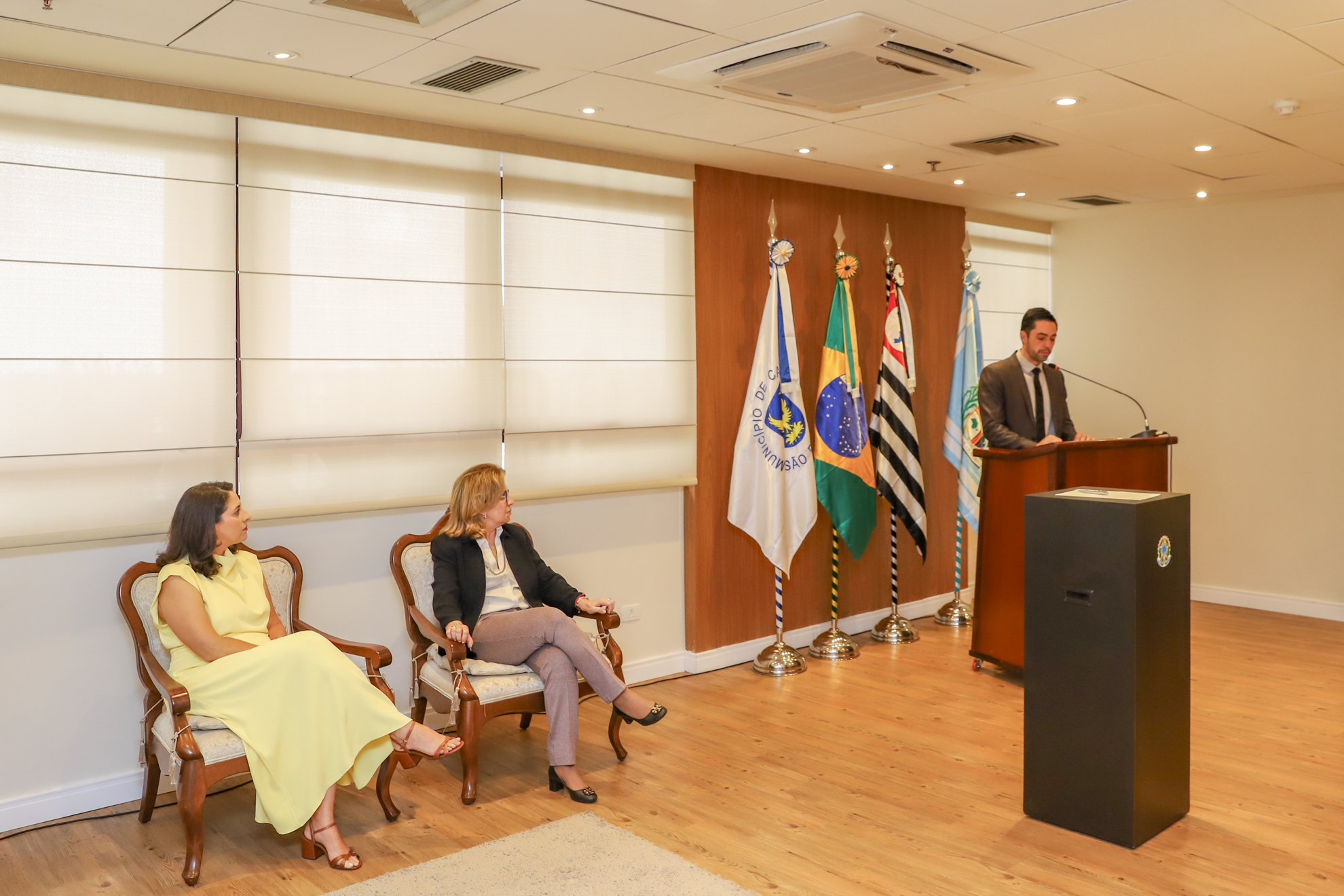 "A juíza Iara e Dra. Ana Paula estão sentadas em cadeiras de madeira, observando o juiz Francisco que está de pé no púlpito falando ao microfone"