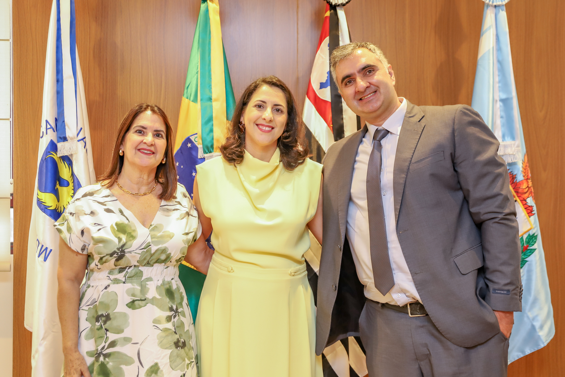 "Dra. Iara posa para foto com a mãe e o marido em frente ao painel de madeira com bandeiras dispostas"