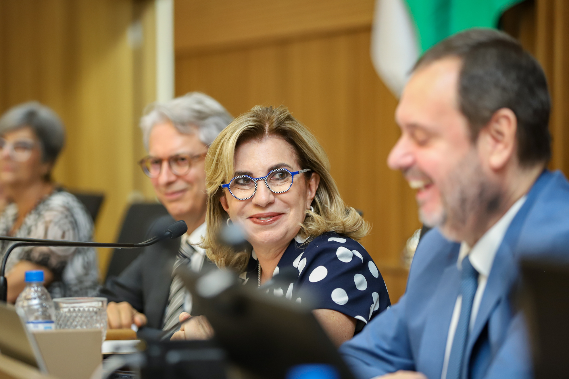 "Dra. Ana Paula, sorridente,  sentada à mesa alta, fala ao microfone, olhando para Paulo Almeida, que está ao seu lado"