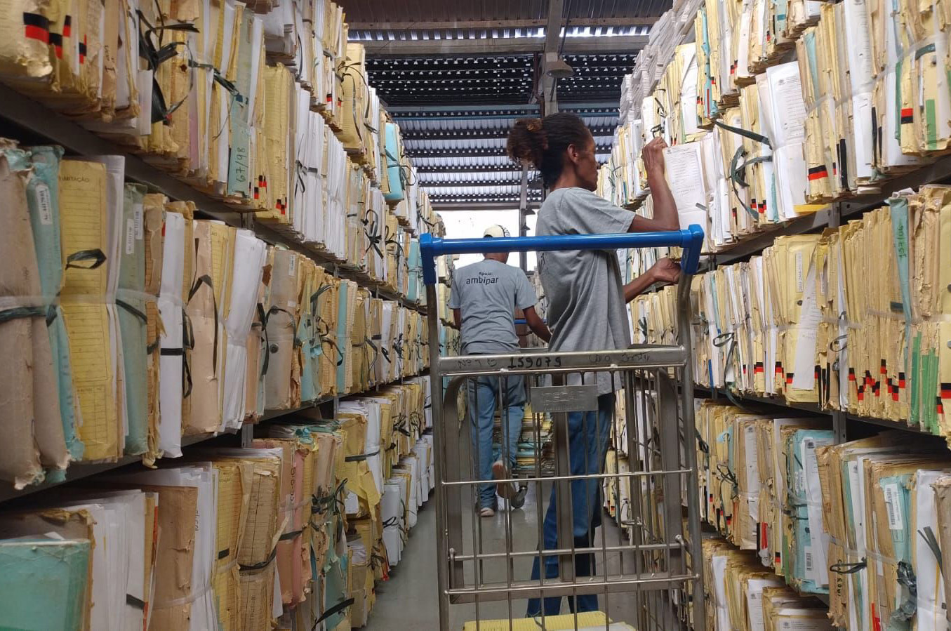 "Duas pessoas da cooperativa retiram processos das estantes para serem levados"