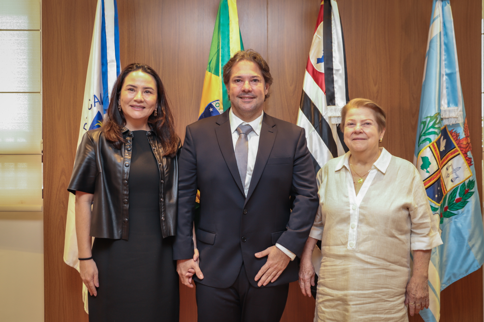 "Dr. Arthur no centro, com a esposa à sua direita, e a mãe, à sua esquerda. Ao fundo, bandeiras em frente a um painel de madeira"