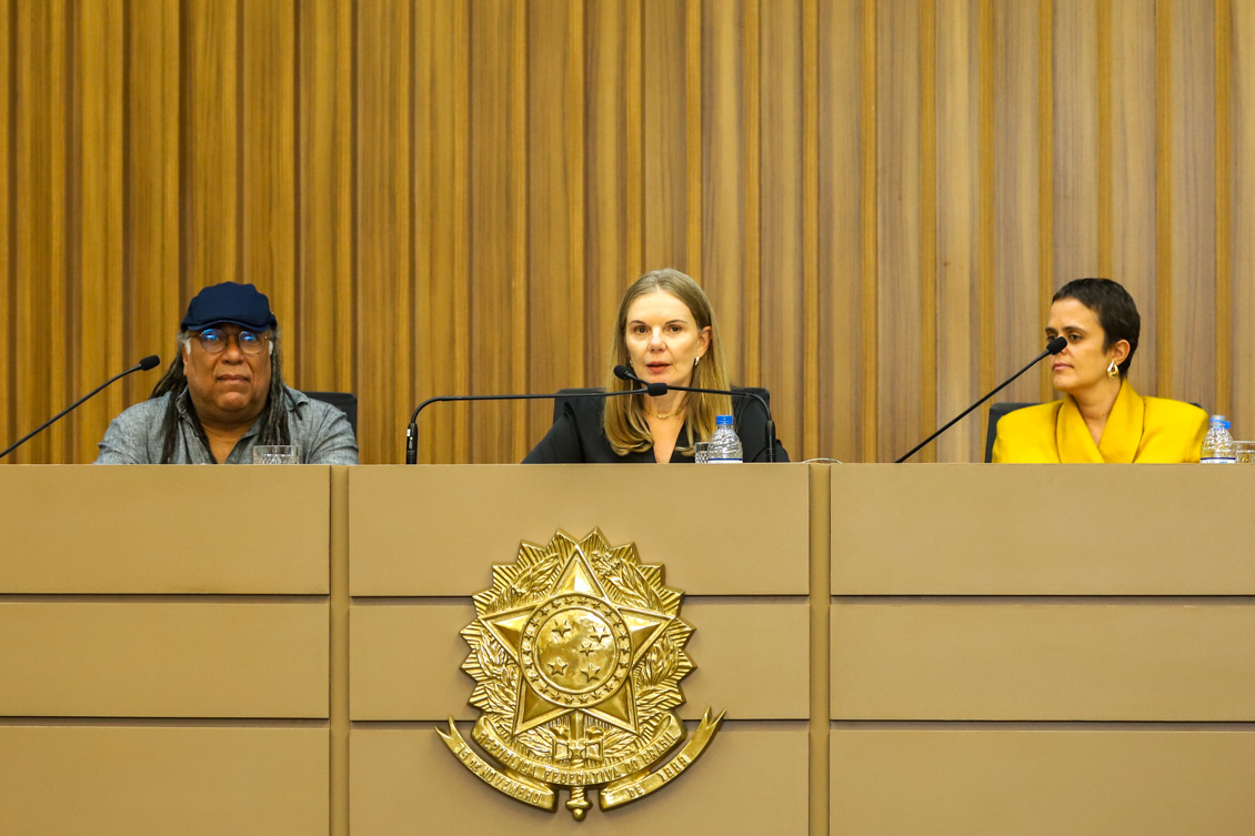 "Desembargadora Maria da Graça comanda painel na mesa alta, entre os dois palestrantes (um homem e uma mulher)"