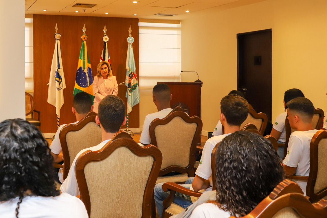 "Dra. Ana Paula está de pé, falando para os aprendizes que estão sentados nas cadeiras do Salão Nobre. Ao fundo, um painel de madeira e quatro bandeiras"
