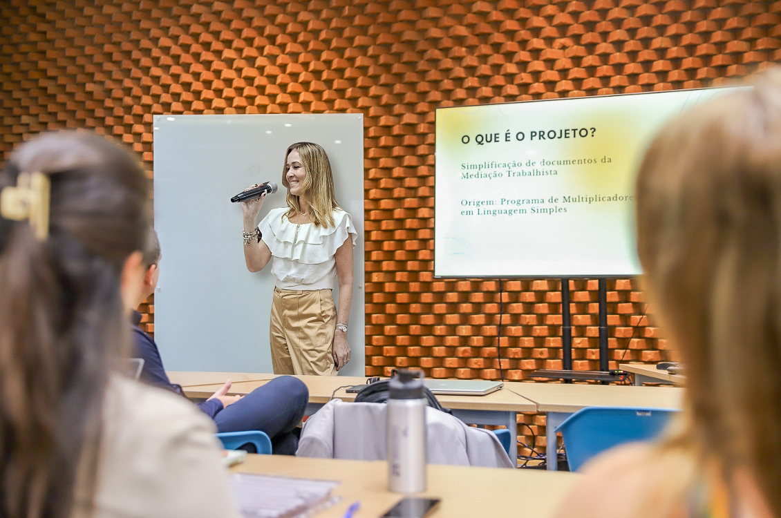 "Dra. Daniela fala ao microfone ao lado de uma tela, sendo observada por participantes"