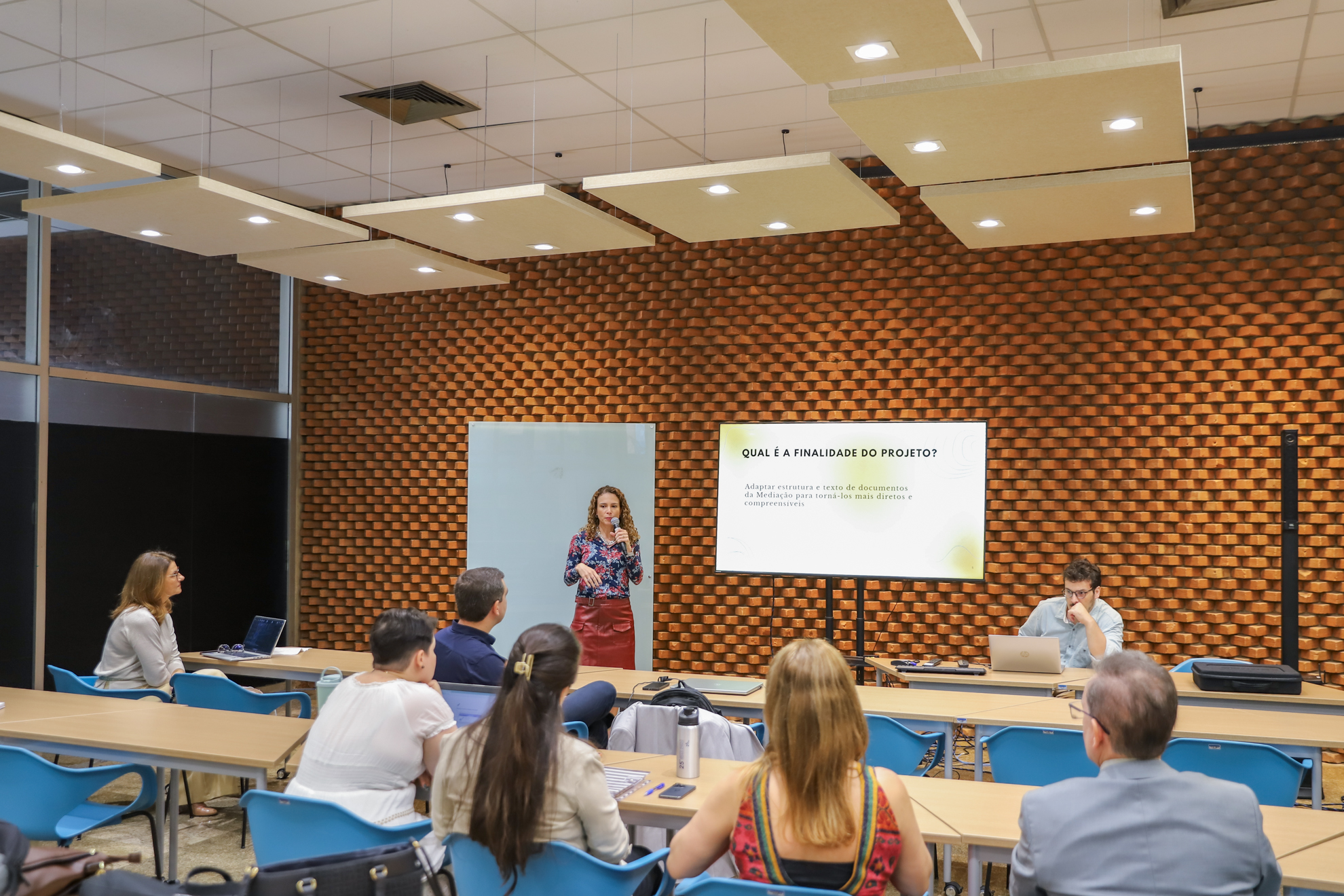 "Dra. Karina fala ao microfone para os participantes, que estão sentados em cadeiras azuis, diante de mesas de madeira clara"