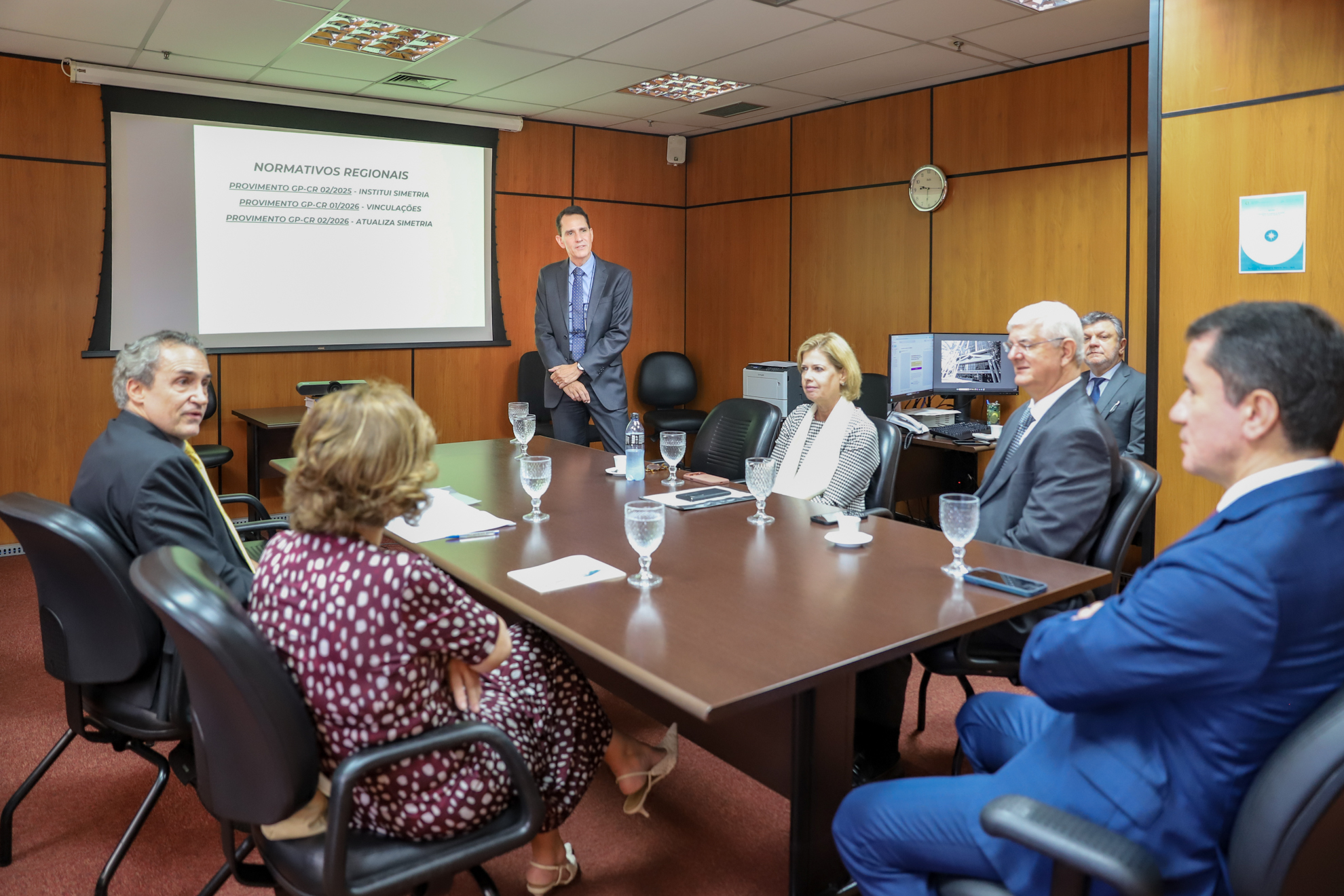 "Dr. Alessandro está de pé ao lado do telão, sendo observado pelos magistrados sentados à mesa"