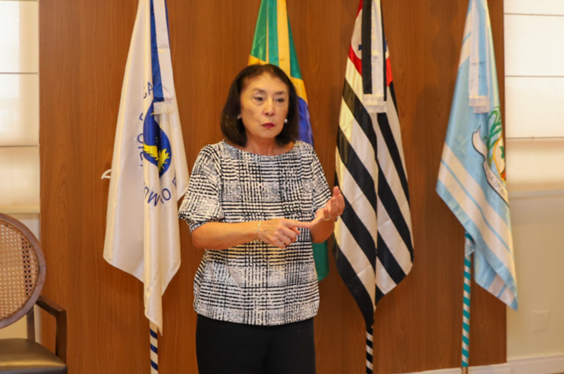 "Sanae, representante do Senac está de pé, em frente ao painel de madeira e bandeiras, gesticulando"