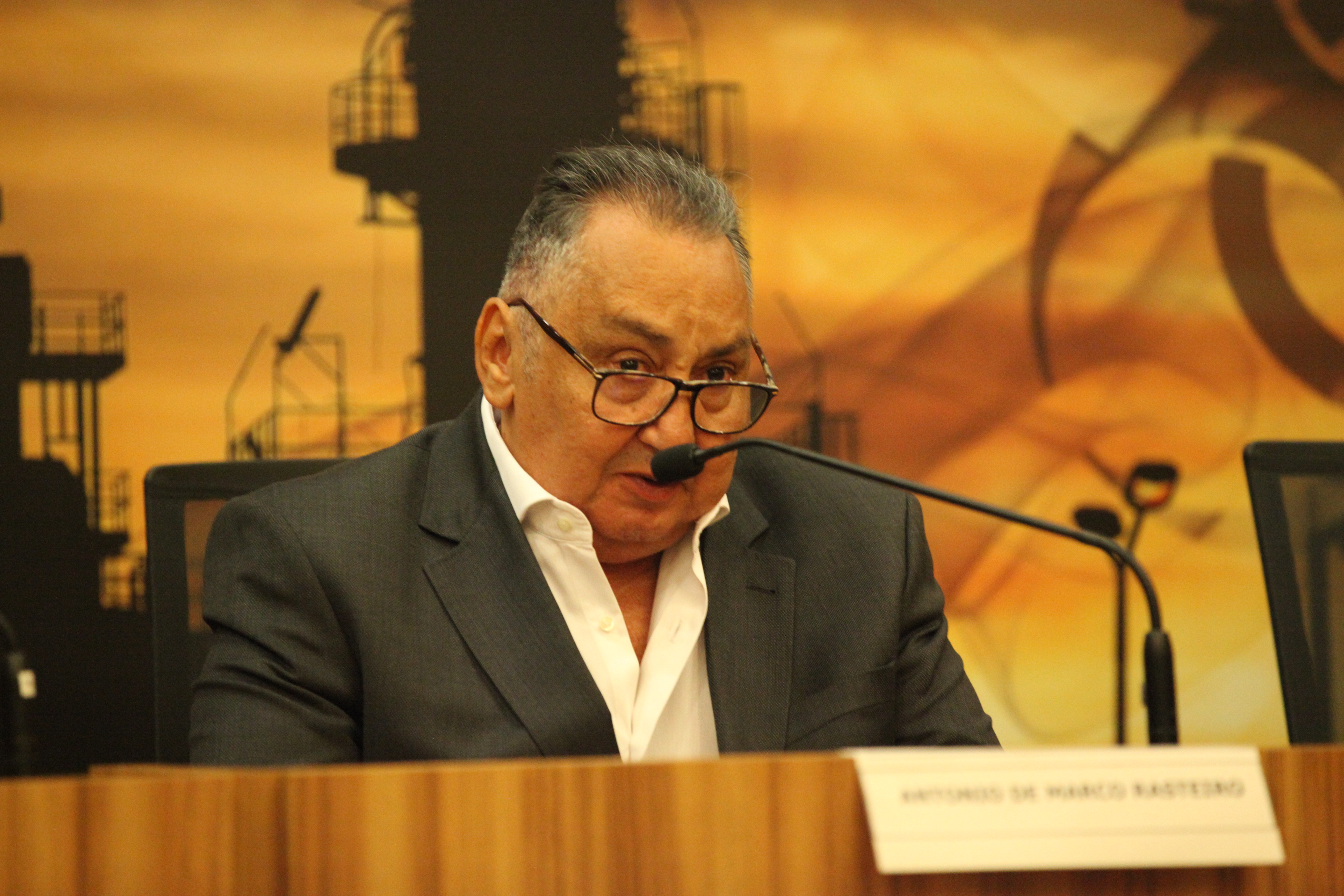 "Antônio Rasteiro fala ao microfone no púlpito com painel ao fundo alusivo ao seminário"