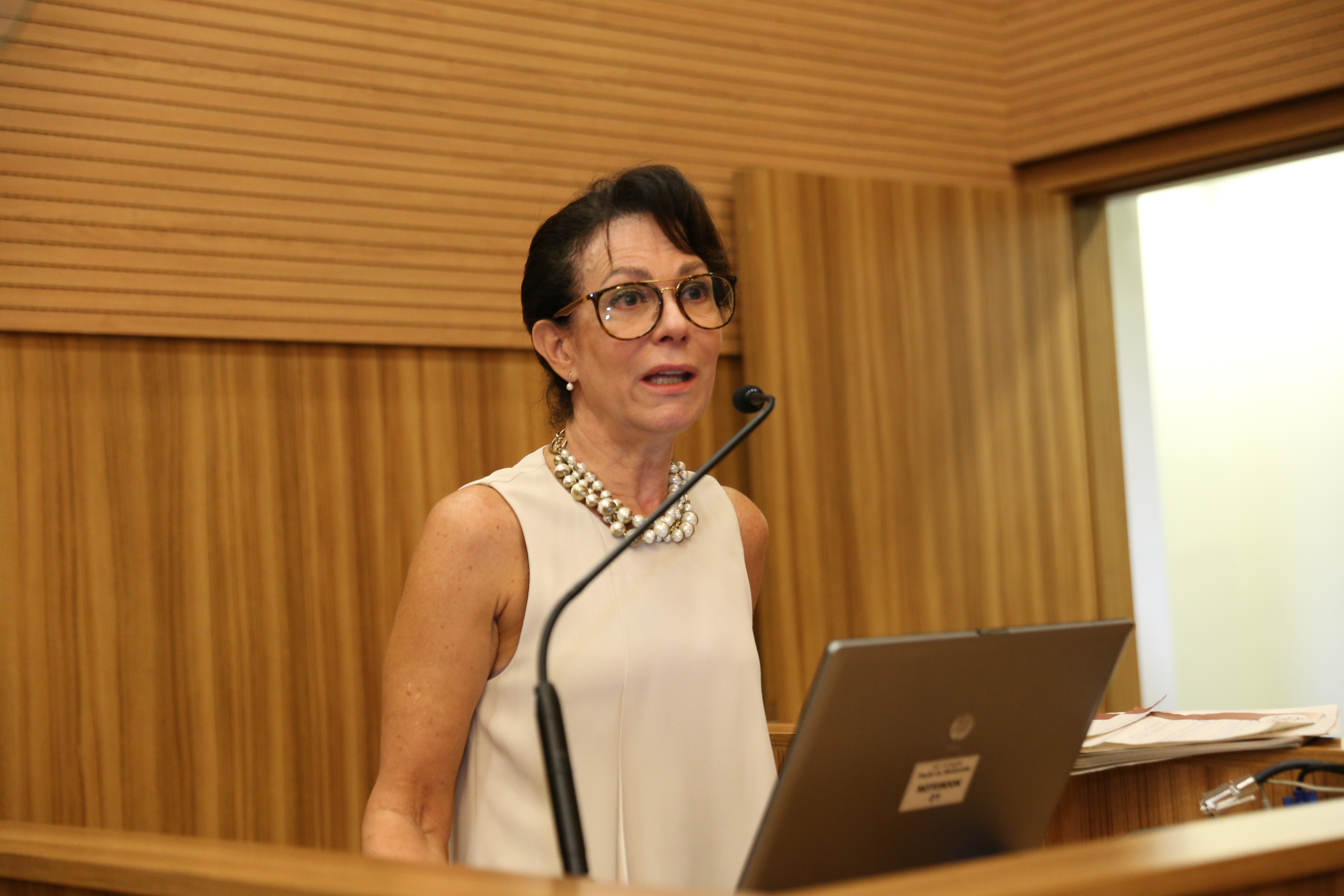 "Dra. Maria Inês fala ao microfone no púlpito com painel de madeira ao fundo"