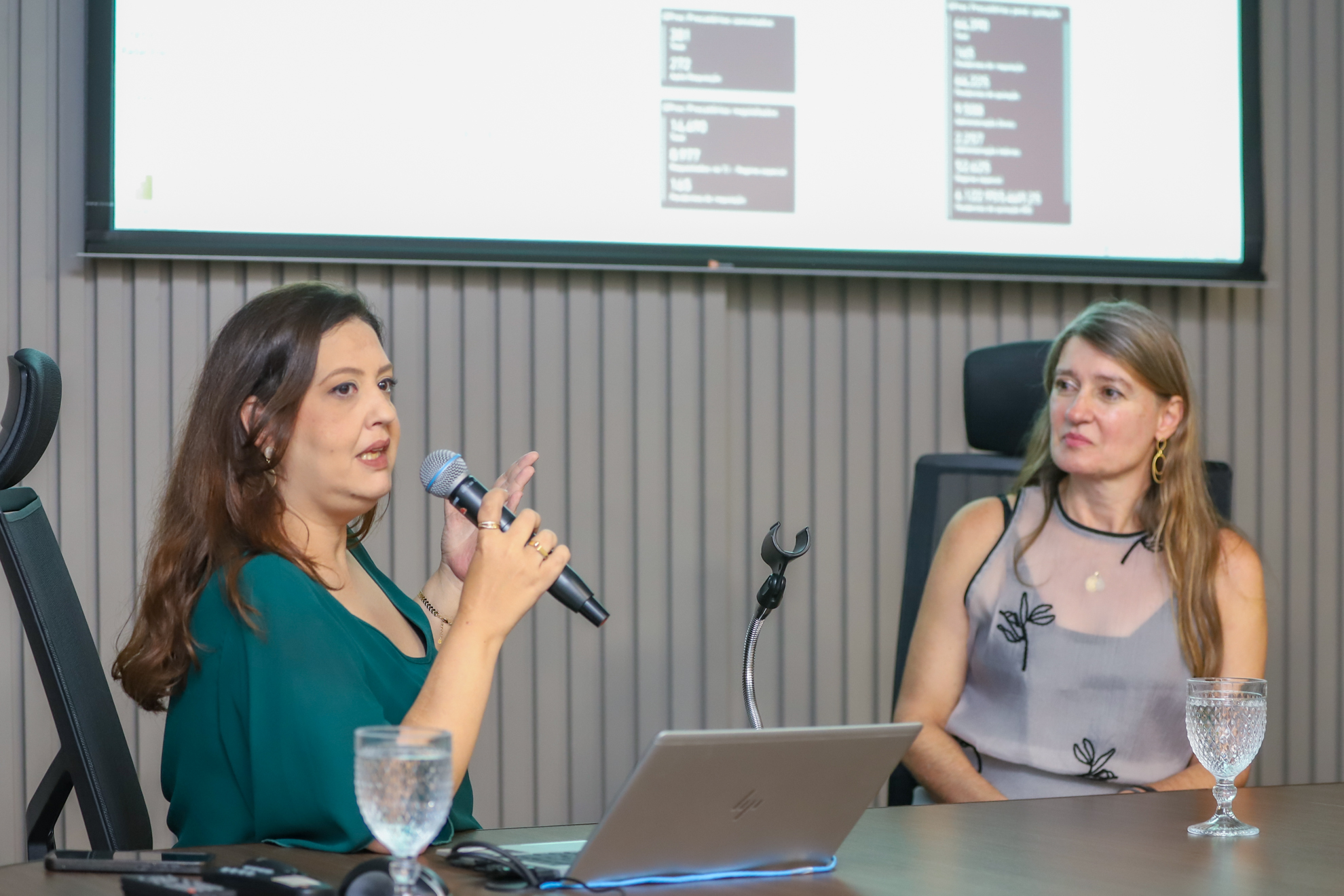 "Servidora Samira, do TRT-15, fala ao microfone apontando para o telão, ao lado de uma outra servidora"