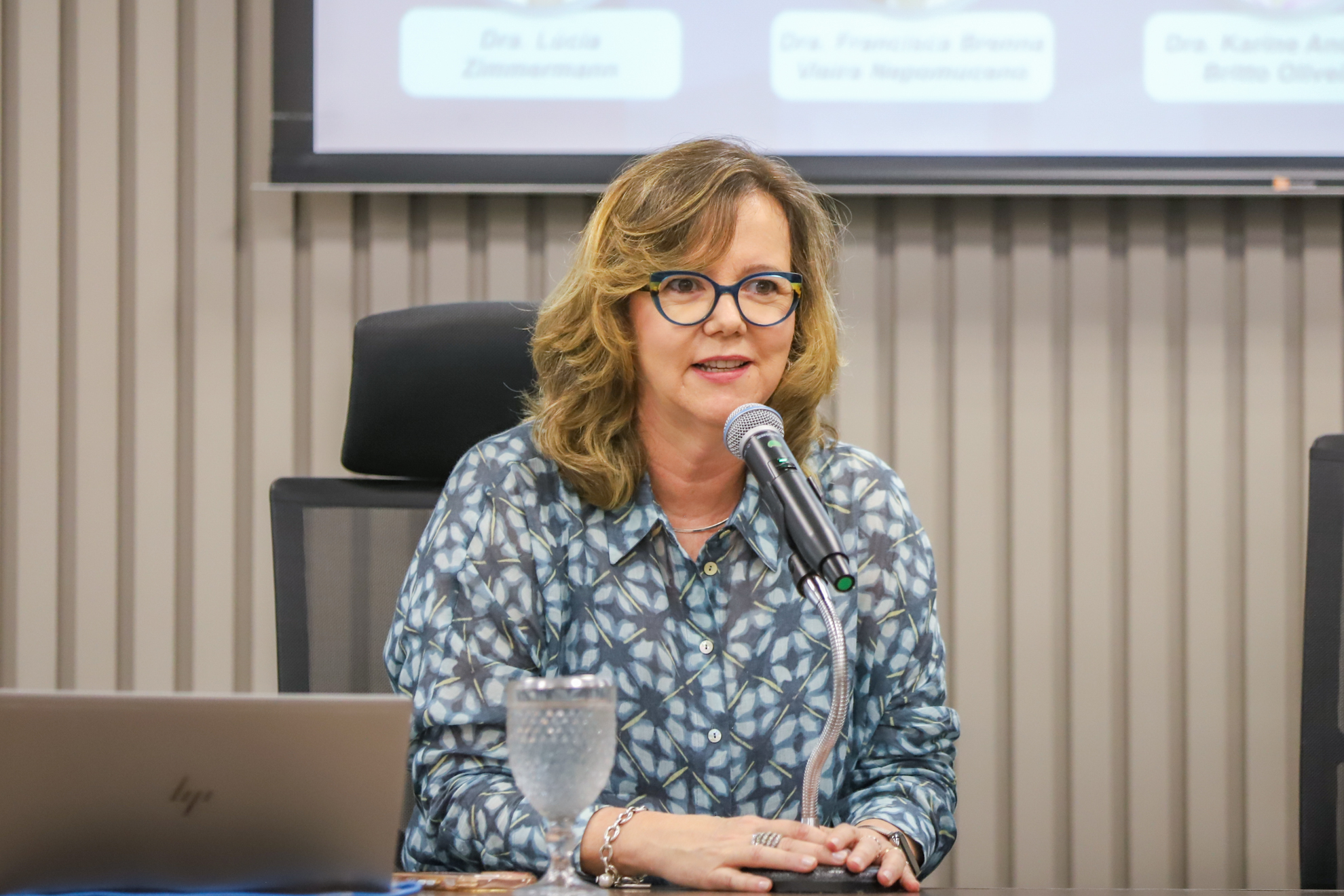"Dra. Eleonora está sentada à mesa alta, falando ao microfone"