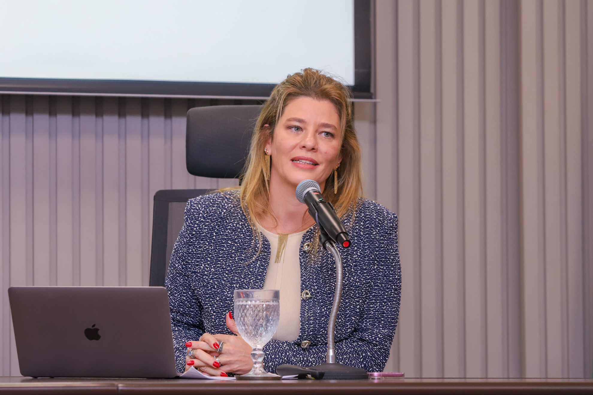 "Dra. Paula está sentada à mesa falando ao microfone, com um notebook da Apple em primeiro plano"