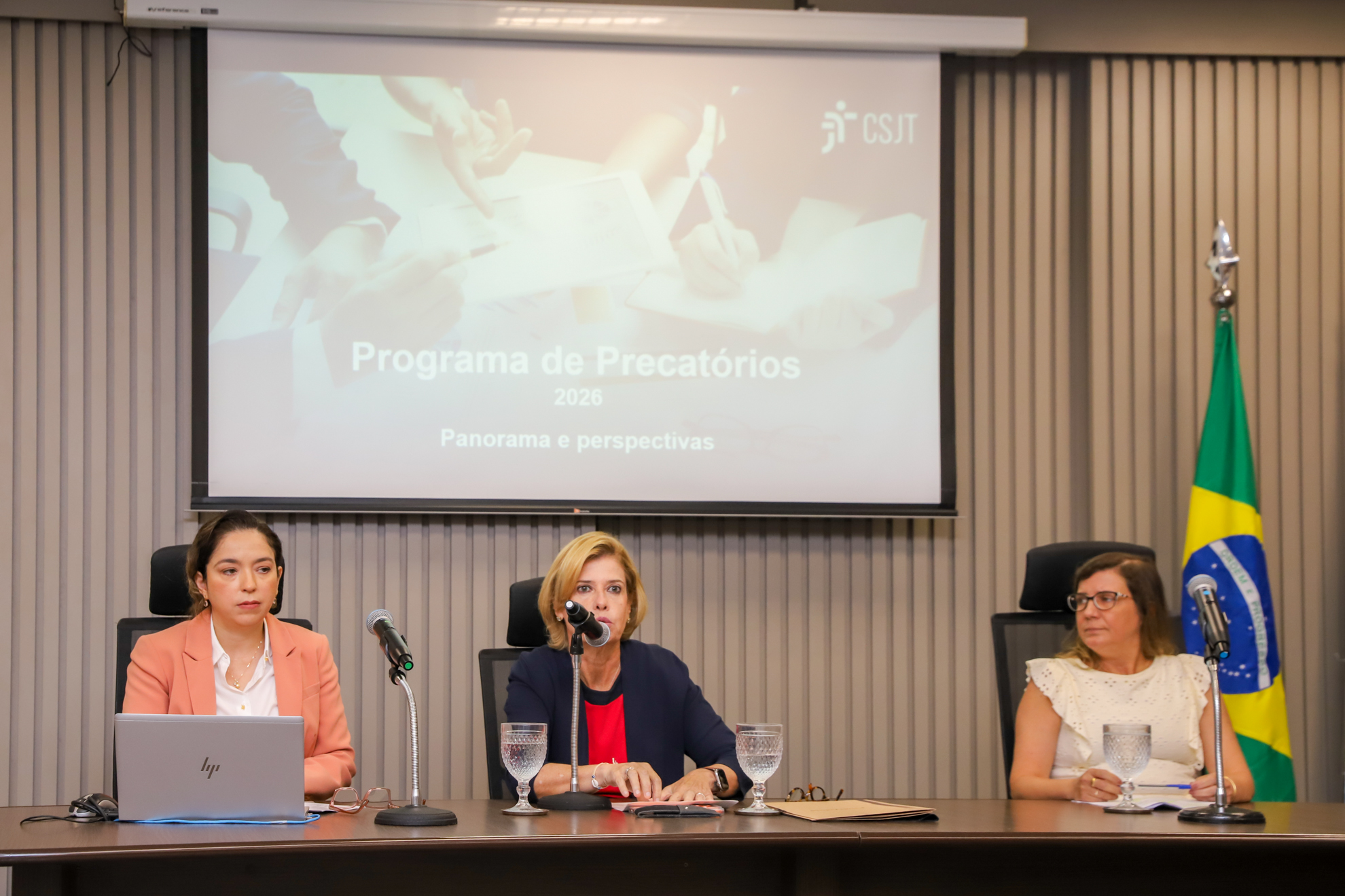 "Dra. Francisca, Dra. Lúcia (ao microfone) e Dra. Karine estão sentadas à mesa de trabalhos, ao fundo o telão temático e a bandeira do Brasil"