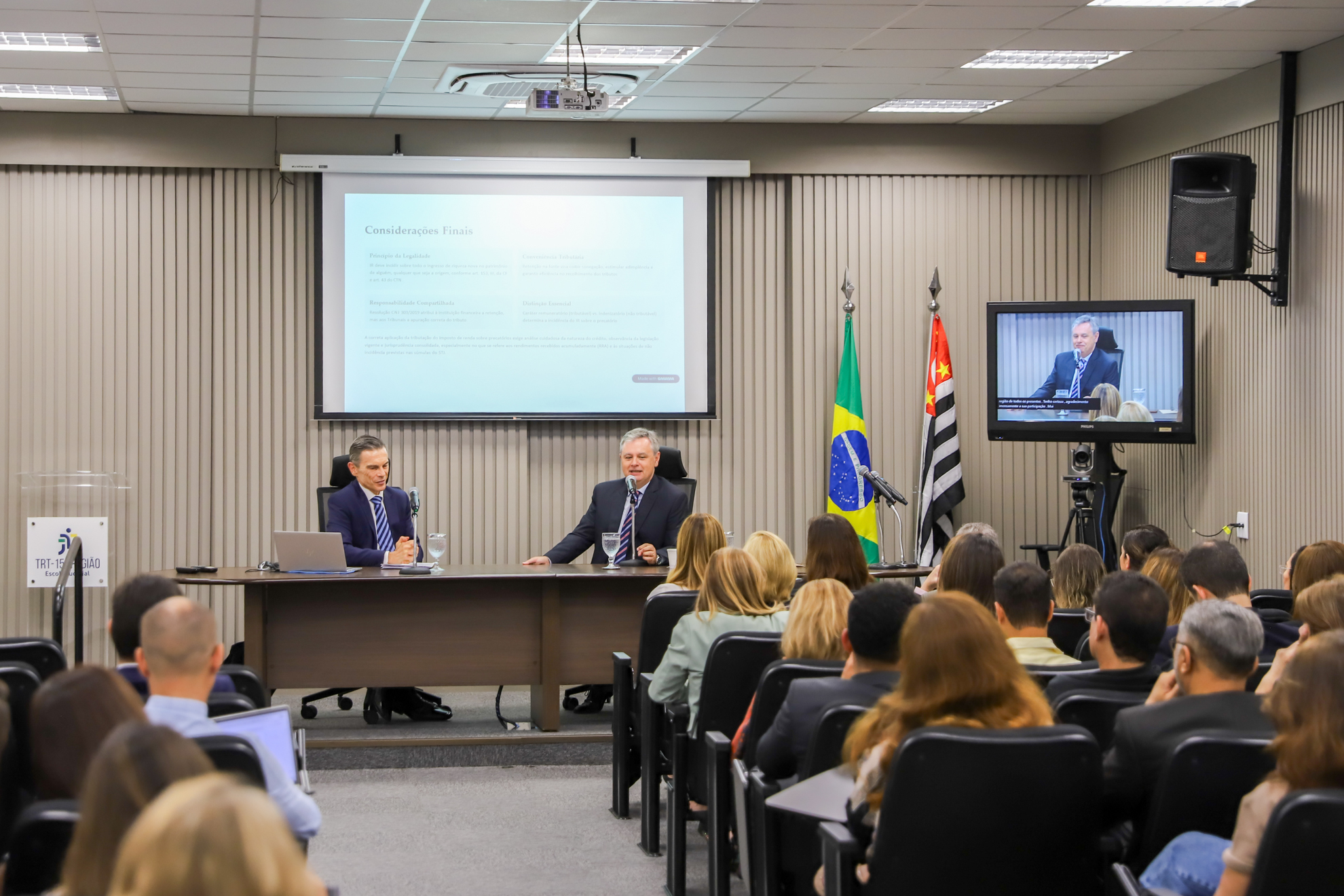 "Foto da mesa de trabalhos ao fundo, composta pelo assessor José Pedro e pelo juiz Flávio Landi, com parte da plateia em primeiro plano"