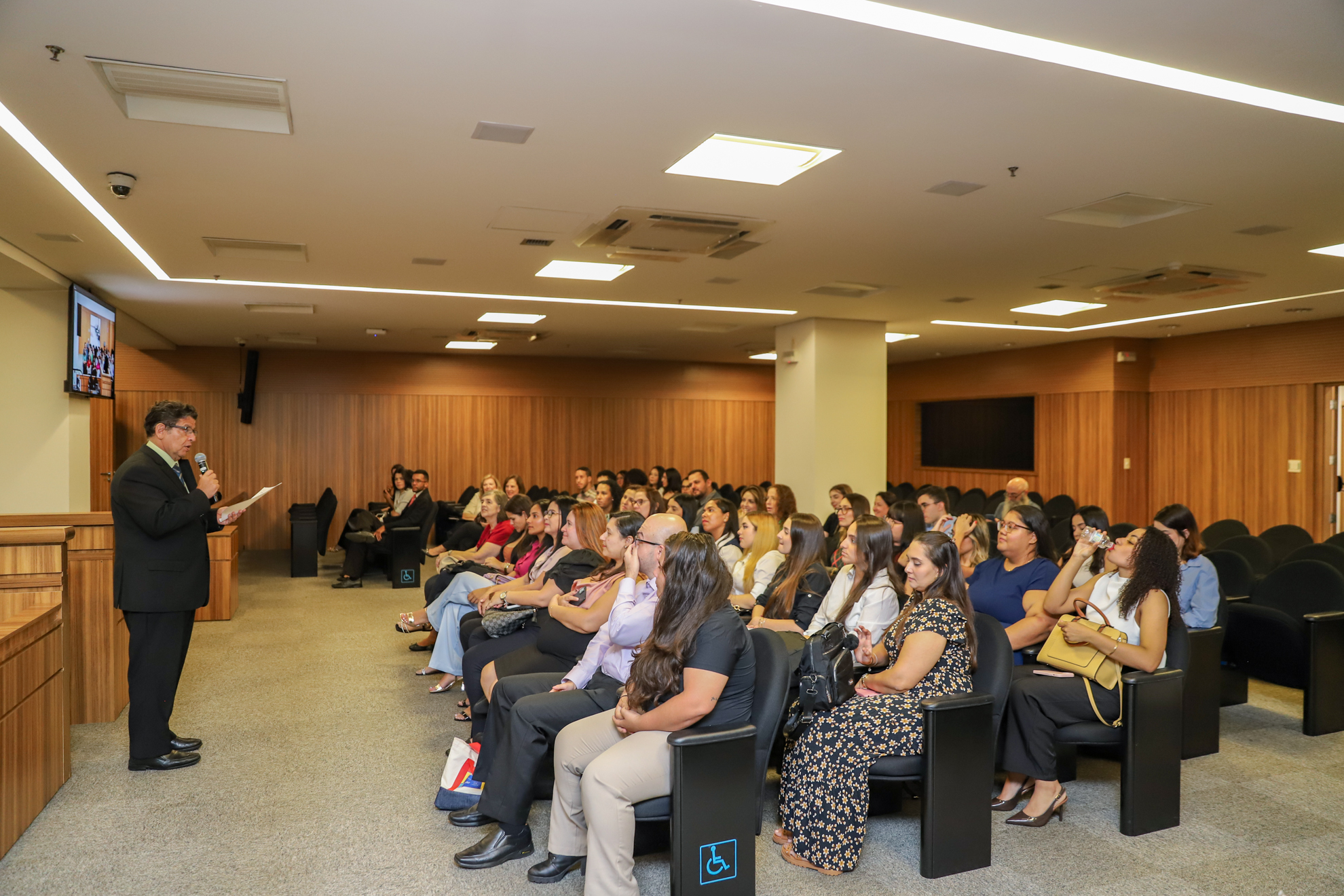 "Dr. Manoel Carlos está com o microfone e papel em mãos, falando aos estudantes, que estão sentados nas cadeiras do plenário""
