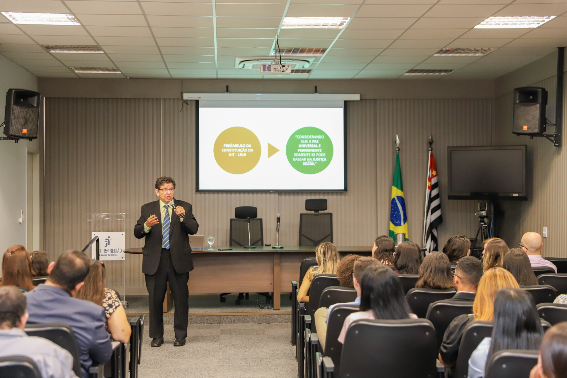 "Dr. Manoel Carlos fala ao microfone no auditório da Ejud15. Ao fundo, um telão e em primeiro plano, os estudantes de costas, sentados em cadeiras"