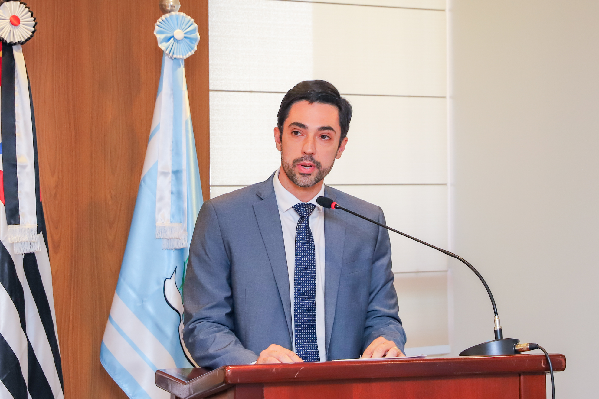 "Juiz Francisco Conte fala ao microfone no púlpito, com as bandeiras ao fundo e painel de madeira"