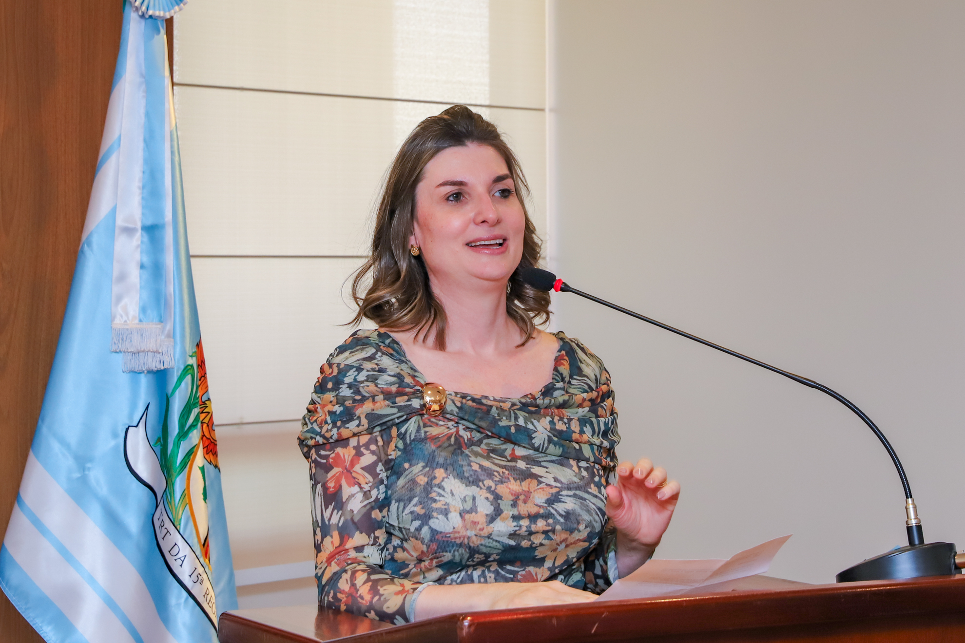 "juíza Andressa fala ao microfone no púlpito com uma bandeira do TRT-15 ao fundo"