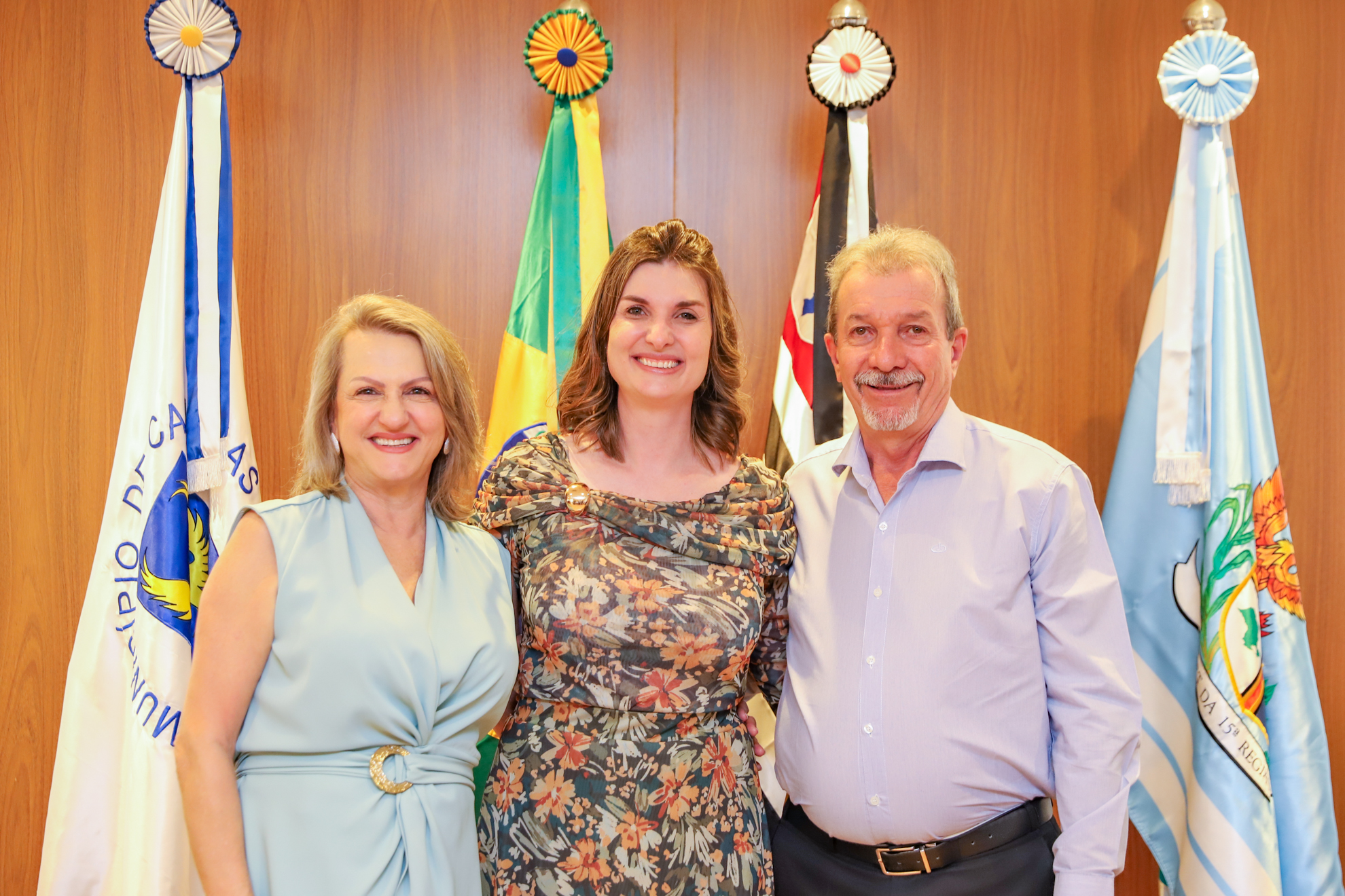 "Juíza Andressa posa para foto com os pais em frente ao painel de madeira e as bandeiras"