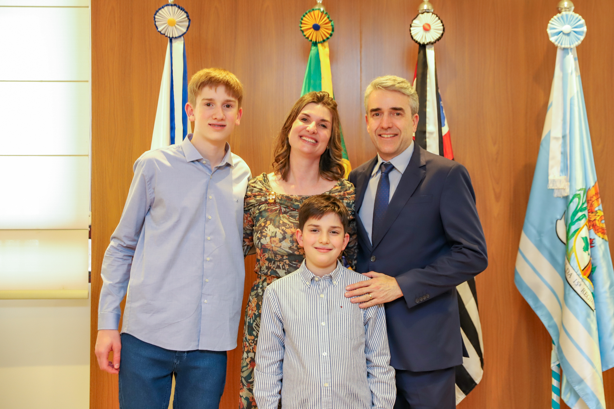 "Juíza Andressa posa para foto com o marido e os dois filhos em frente ao painel de madeira e as bandeiras"