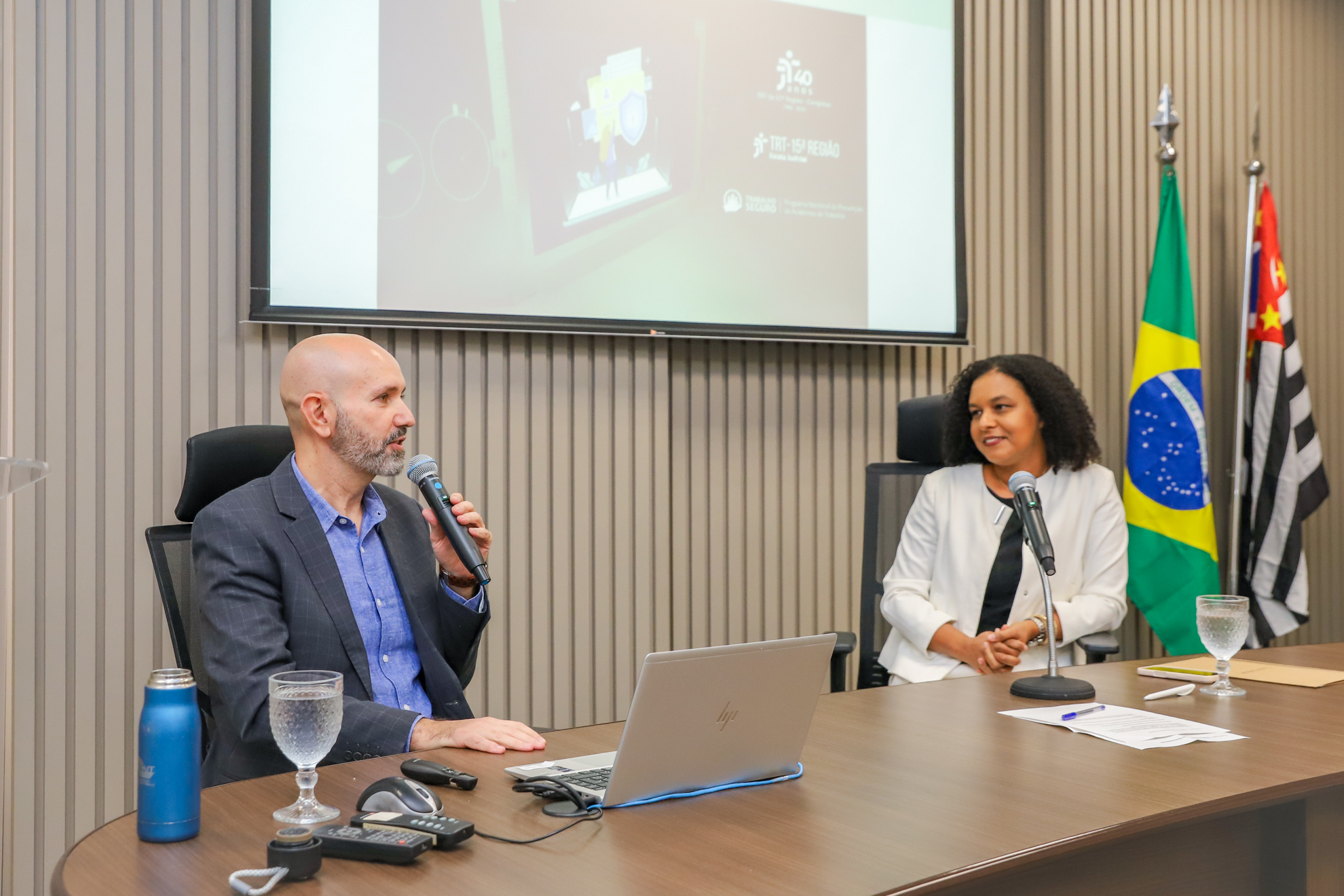 "#ParaTodosVerem: auditor Rodrigo e juíza Paula estão sentados atrás da mesa de trabalhos do auditório da Ejud15"