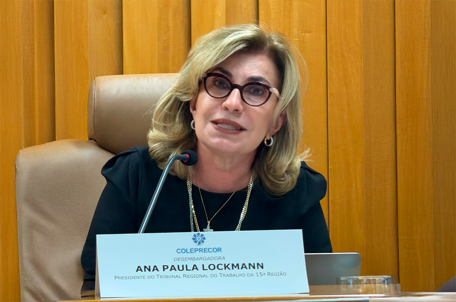 "Dra. Ana Paula fala ao microfone à mesa, que exibe placa de identificação com logotipo do Coleprecor"