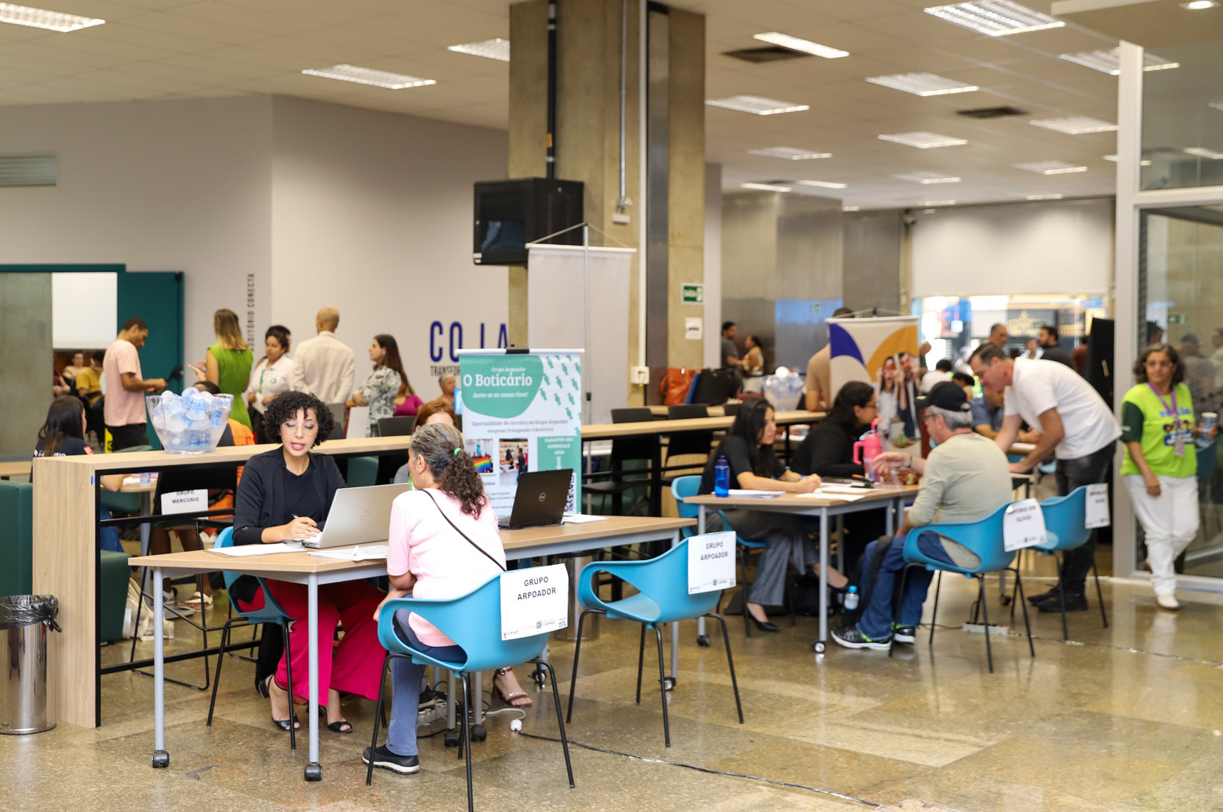 "Foto geral do evento com pessoas sendo atendidas em mesas, outros ao fundo, em pé"